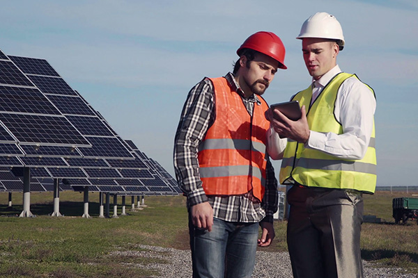 Sustainability and energy team working. Two men in hard hats and vests looking at a tablet in front of solar panels.